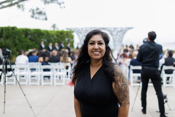 Woman with long hair, wearing a black blouse smiling in front of a wedding event.